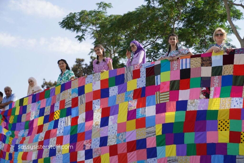 A super large quilt made by many woman who are all holding up the quilt from a stand.