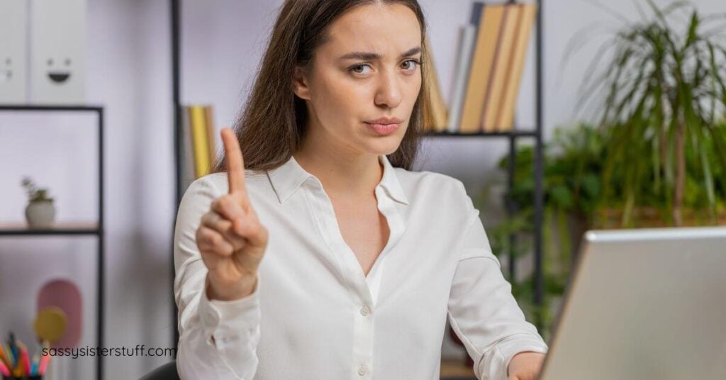 midlife woman sitting at a desk holds up on finger to let a visitor know she's busy.