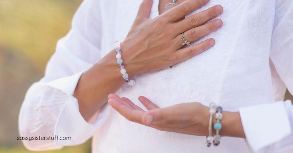 close-up of woman in white shirt with one hand to her heart and one hand facing up toward the sky.