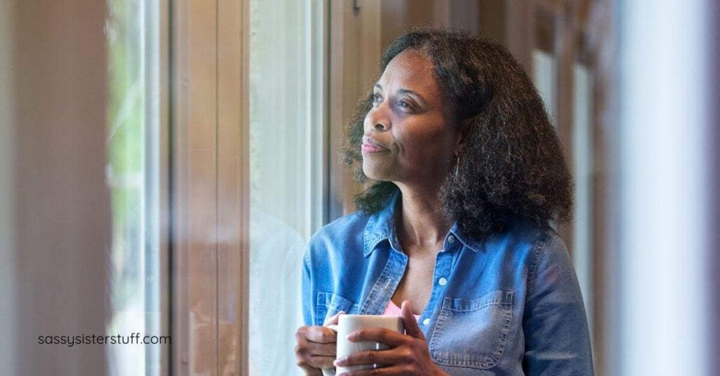 midlife woman with a cup of tea look out a window up toward the sky as she thinks about the power of reflection and finding her north star.