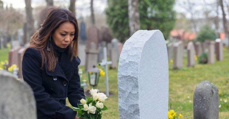 a woman sadly sits graveside with white flowers for a loved one.
