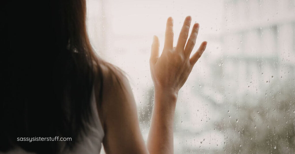 woman with her hand up to a window as she looks out on a rainy day and reflects on her life.