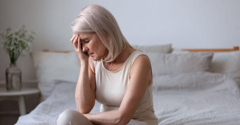 middle age woman sitting on her bed with her hand to her forehead in despair.