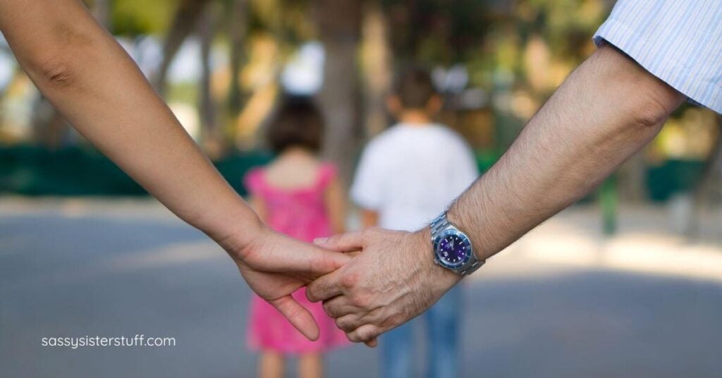 mother and father hold hands in a photo with their two children blurred in the background between their parents' hands.