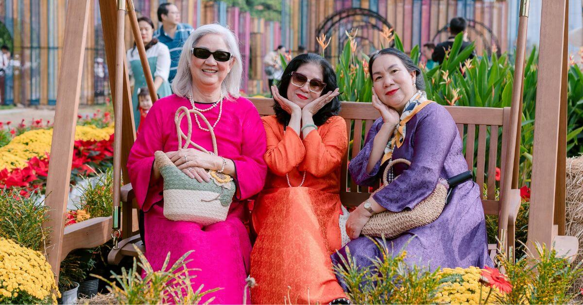 three happy ladies in bright colors sit on a wooden swing among colorful flowers.