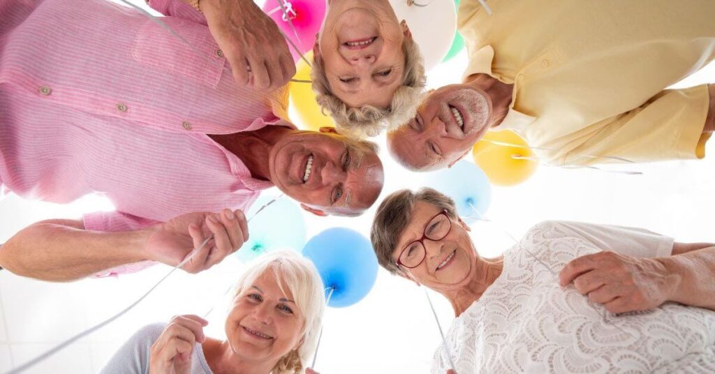 close up of five elderly and midlife friends holding balloons in a circle looking down at a camera.