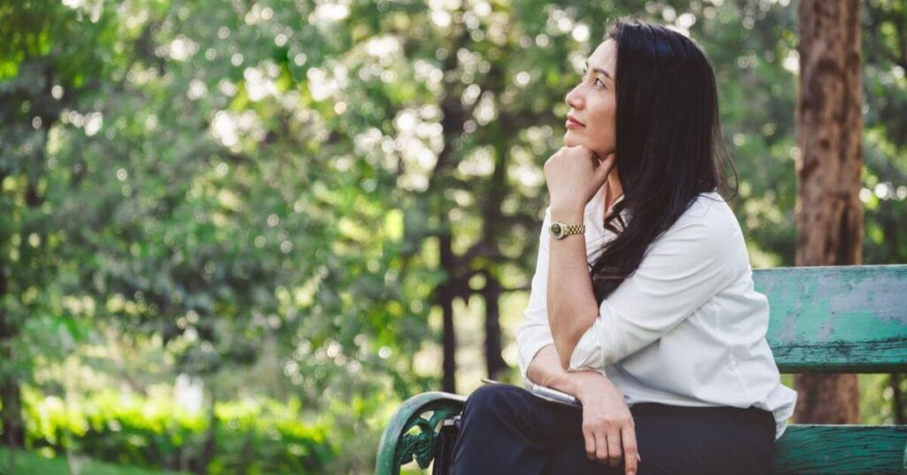 Asian woman sits on a bench in the park planning the perfect reset day.