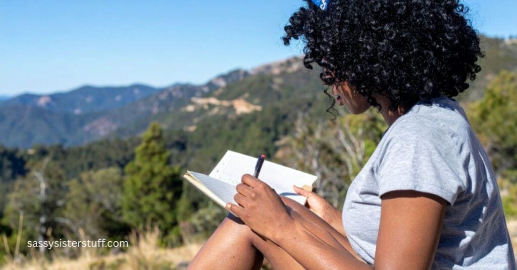 woman sitting on a hill in the mountains writing in her journal.
