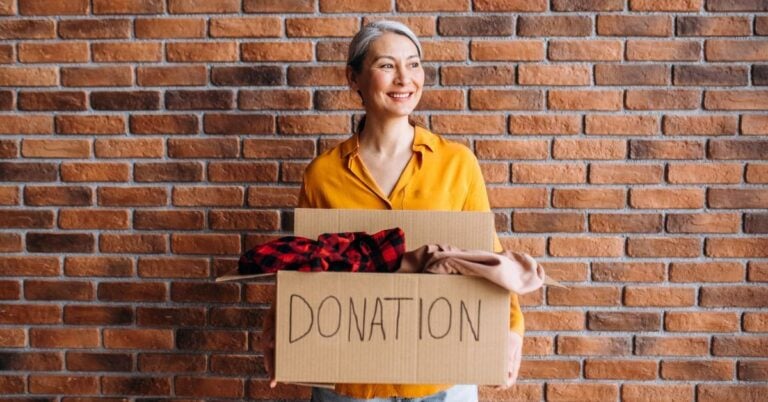 middle aged woman stands against a brick wall holding a box marked DONATION with a smile on her face because she is supporting a local cause.