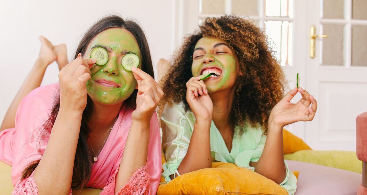 two women enjoy a facial mask and cucumbers as part of their self-care habits for midlife women while laying on a bed laughing.