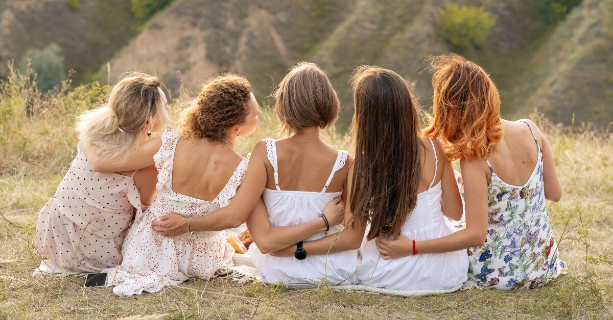 five women in delicate sundresses sitting on a hill with their arms around each other.