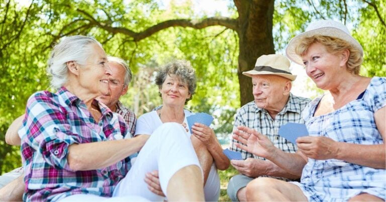 a group of senior friends sit together in a park and play cards.