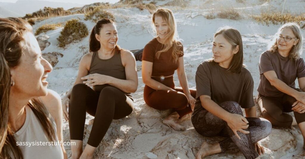 a group of 5 women who are working to build a support system for each other sit on a sandy beach and chat.
