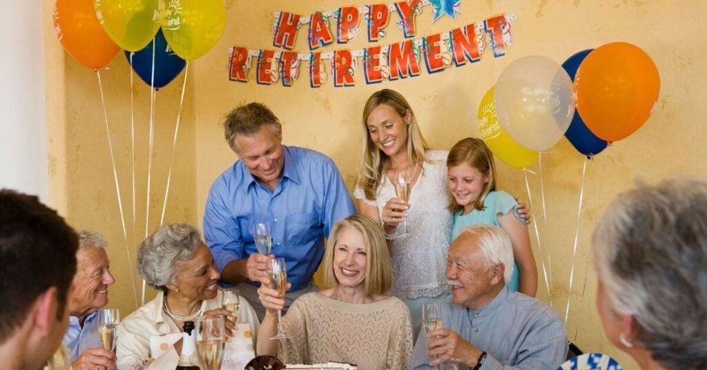 A group of adults and a child raising glasses around a cake at a table with balloons and retirement signs.