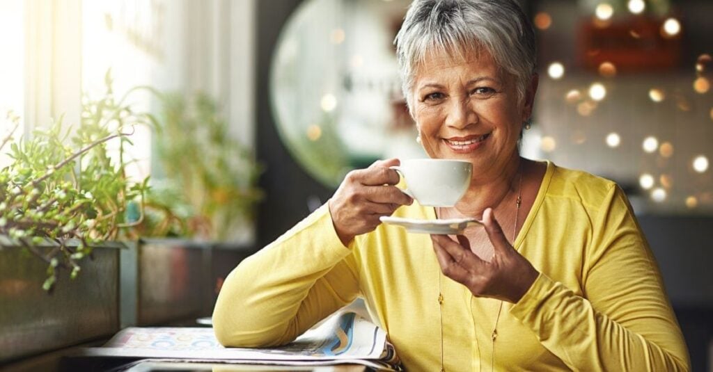 A smiling older woman wearing a yellow blouse lifts a white coffee cup while sitting alone at a café.