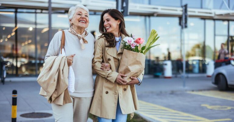 two happy older women walk out of a grocery store with a bag of flowers and healthy food.